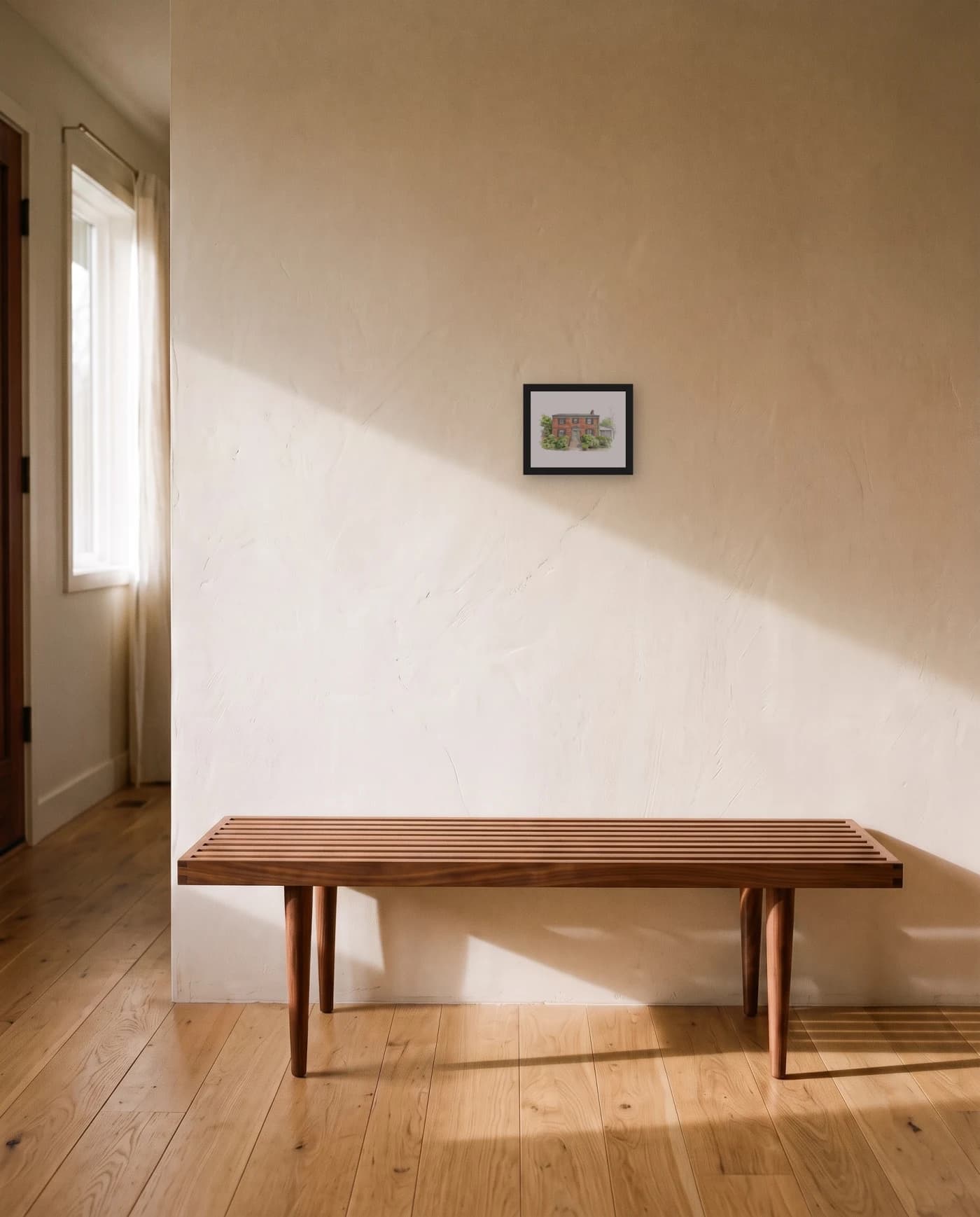 A framed house portrait in a black frame on a plaster entry wall, above a low walnut slat bench in morning light.