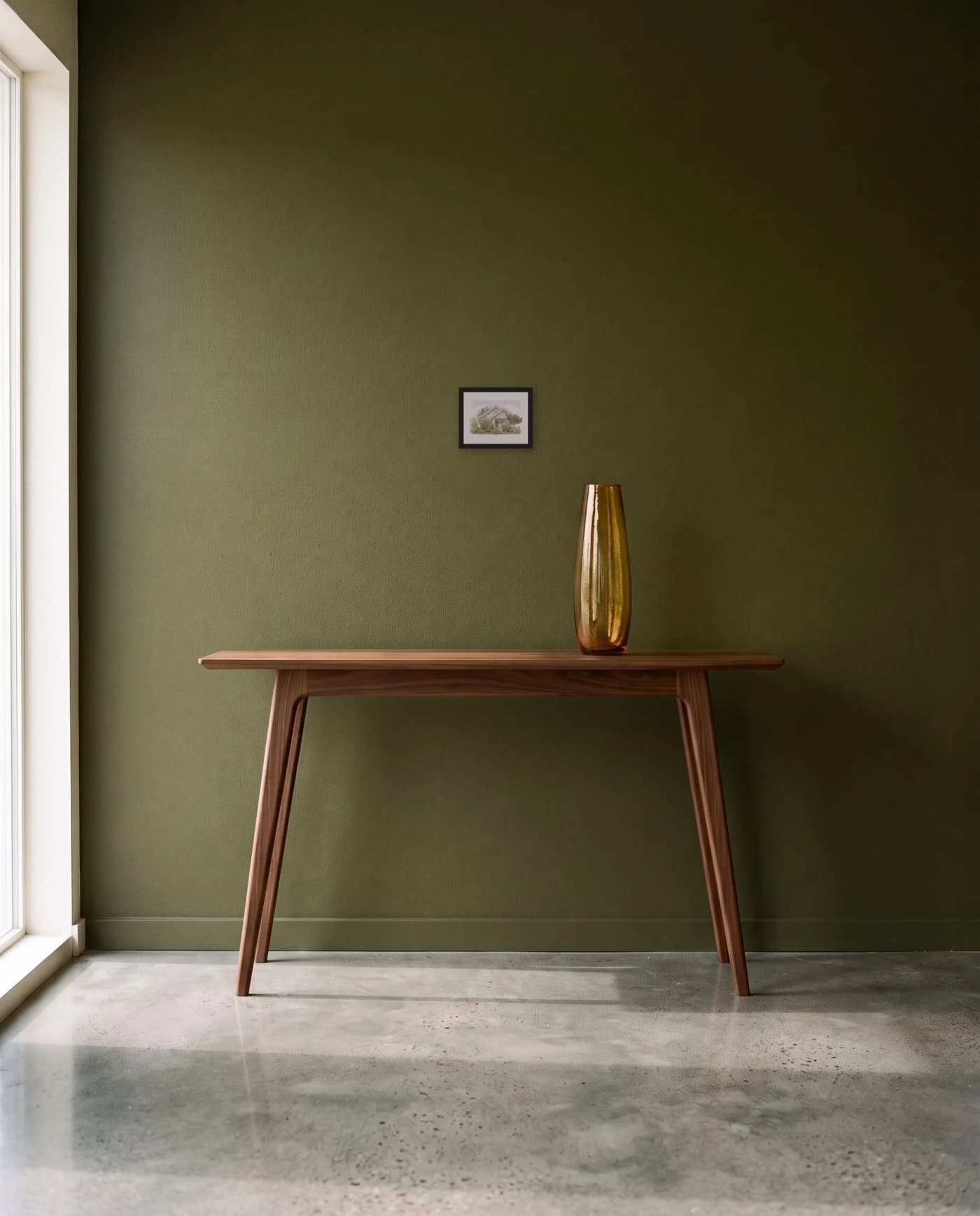 A framed house portrait in a black frame on a deep olive wall, above a slim walnut console with a tall amber glass vessel.