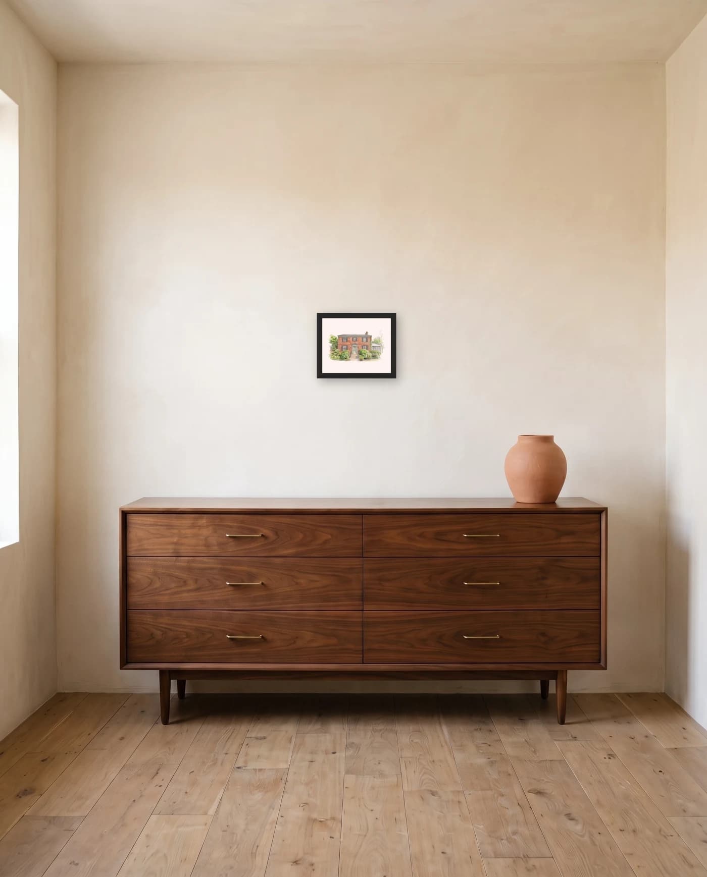 A framed house portrait in a black frame above a long low walnut dresser, one terracotta vessel at the far end.