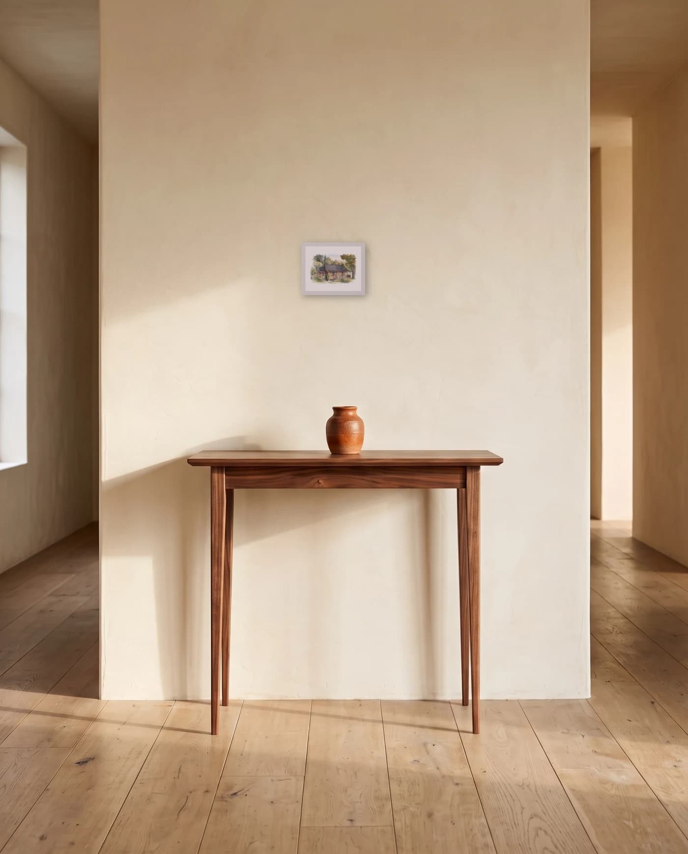 A framed house portrait in a white frame on a plaster hallway wall, above a narrow walnut table with a single ceramic vessel.
