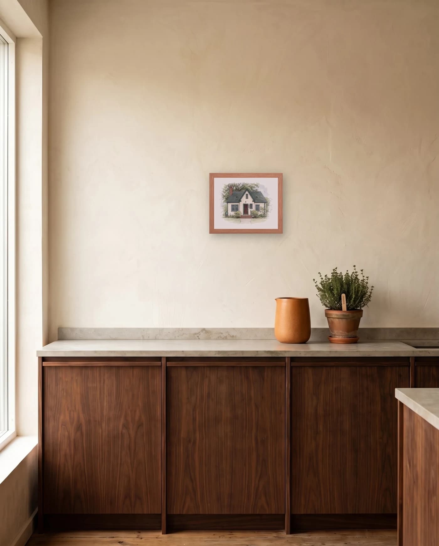 A framed house portrait in a red oak frame on a plaster kitchen wall, above a walnut counter run with a ceramic vessel and a potted herb.