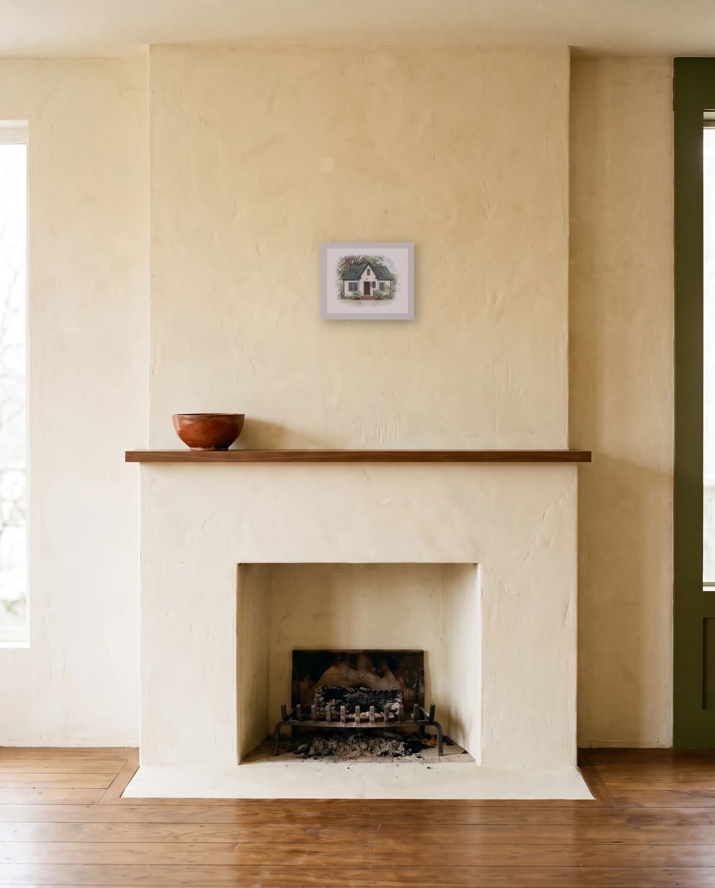 A framed house portrait in a white frame above a plain plastered mantel shelf holding one low ceramic bowl.