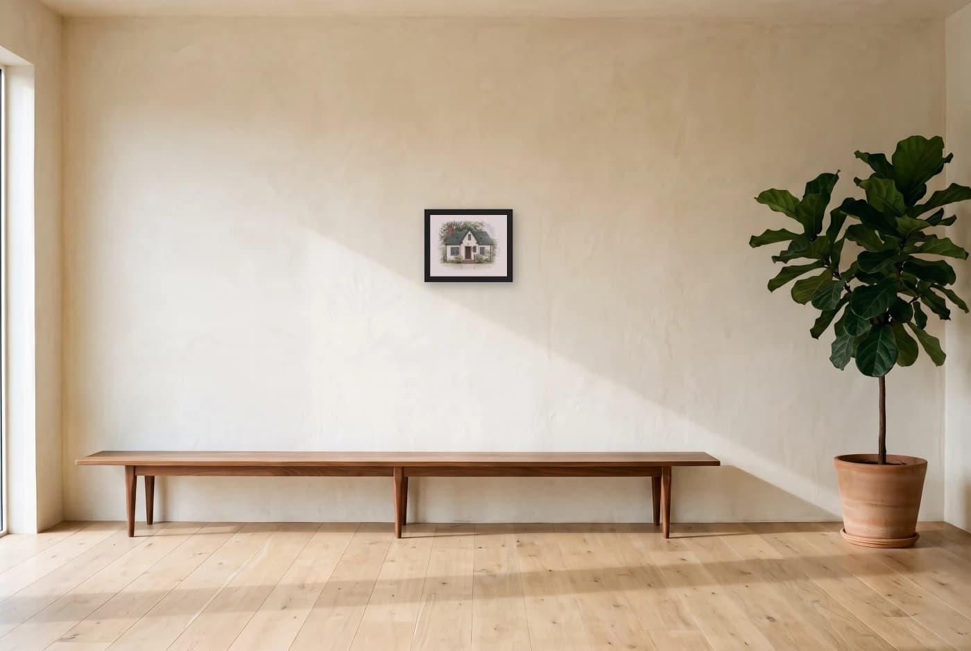A framed house portrait in a black frame on a wide plaster wall, a low walnut bench along it and a potted fig at the edge.
